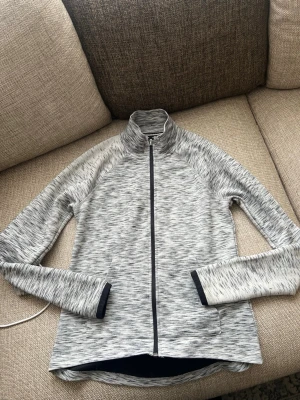Gråmelerad ziptröja från Forever 21 - Sportig gråmelerad ziptröja från Forever 21 med hög krage och hel dragkedja framtill. Tröjan har långa ärmar och tumhål vid ärmsluten, perfekt för träning eller chill. Materialet är mjukt och stretchigt för extra komfort.