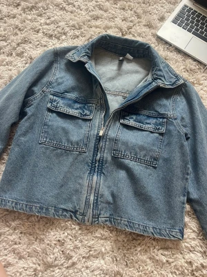 Blå jeansjacka från H&M med dragkedja - Snygg blå jeansjacka från H&M med dragkedja framtill och två stora bröstfickor med lock. Klassisk krage och raka ärmar. Perfekt att slänga över en hoodie eller t-shirt för en avslappnad look.