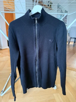 Svart ribbad ziptröja från Puma - Svart ribbstickad ziptröja från Puma med hög krage och diskret logga på bröstet. Tröjan har hel dragkedja framtill och är tillverkad i ett mjukt, stretchigt material. Perfekt för en sportig och stilren look.