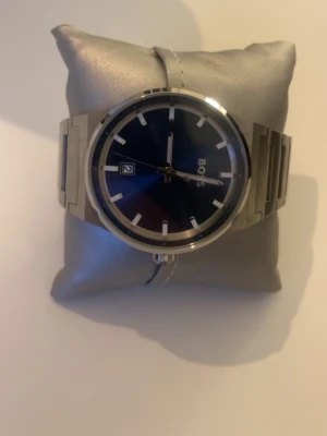 Silverfärgad klocka från BOSS - Stilren klocka från BOSS med silverfärgat armband i metall och mörkblå urtavla. Klockan har tydliga indexmarkeringar, datumvisning och klassisk rund form. Perfekt accessoar för dig som gillar en clean och modern look.