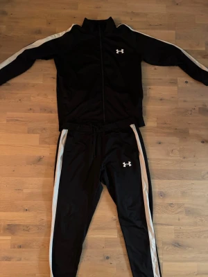 Svart träningsset från Under Armour - Snyggt svart träningsset från Under Armour med vita ränder längs ärmar och ben. Setet består av en zip-jacka och matchande byxor, båda med logga på bröst och lår. Perfekt för gymmet eller sportaktiviteter.