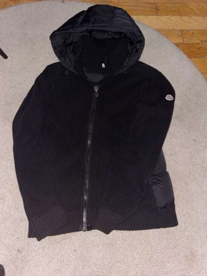 Svart pufferjacka från Moncler - Svart pufferjacka från Moncler med stickade ärmar och vadderad kropp. Jackan har huva, dragkedja framtill och Moncler-logga på ärmen. Perfekt för kalla dagar med sin kombination av dun och stickat material.