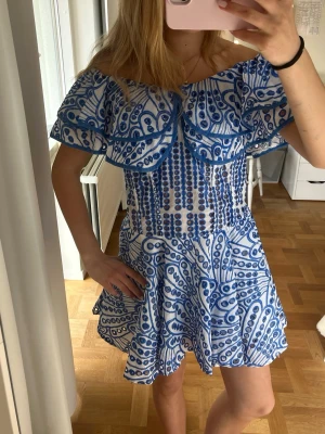 Blåvit offshoulder kortklänning - Superfin blåvit kortklänning med offshoulder och volangdetaljer. Klänningen har ett unikt mönster i blått på vit botten och en luftig, snurrig kjol. Perfekt för varma dagar och sommarens alla tillfällen.