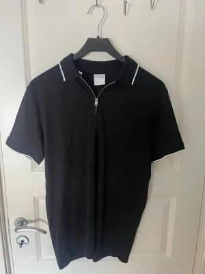 Svart piké med half zip från Selected - Snygg svart piké från Selected med vit piping på kragen och ärmsluten. Tröjan har korta ärmar och en stilren half zip-dragkedja framtill. Tillverkad i mjuk bomull för skön känsla och enkel stil.
