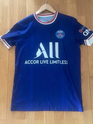PSG matchtröja Mbappé #7 blå - Snygg blå Paris Saint-Germain matchtröja med Mbappé och nummer 7 på ryggen. Tröjan har vita och röda detaljer vid krage och ärmslut, samt klubbmärke och sponsortryck framtill. Perfekt för fotbollsfans som vill sticka ut på planen eller läktaren.