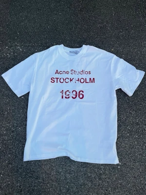 Vit Acne Studios t-shirt Stockholm - Säljer en vit t-shirt från Acne Studios med röd text 'STOCKHOLM 1996' framtill. Klassisk passform och rund halsringning. Perfekt basic med cool print för dig som gillar stilrena plagg med en twist.