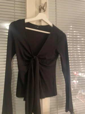 Svart omlottopp med knyt från Gina Tricot - Snygg svart omlottopp från Gina Tricot med långa ärmar och knyt framtill. Toppen har en djup v-ringning och är tillverkad i ett mjukt, stretchigt material som sitter skönt på kroppen. Perfekt att styla med jeans eller kjol.