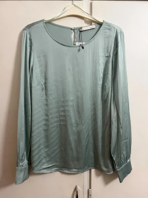 Ljusgrön satinblus från Oui - Snygg ljusgrön blus i satin från Oui med rund halsringning och långa ärmar. Blusen har breda manschetter och en knapp i nacken för stängning. Perfekt för en stilren och trendig look.