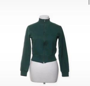 Zara trackjacka - Stilren mörkgrön jacka med hög krage och hel dragkedja framtill. Modellen är kort och har bred mudd i midjan samt vid ärmsluten. Perfekt för dig som gillar en sportig och enkel look.