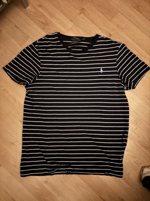 Randig t-shirt från Polo Ralph Lauren - Svart t-shirt med vita horisontella ränder från Polo Ralph Lauren. Klassisk rund halsringning och broderad logga på bröstet. Modellen är slim fit och passar perfekt till en avslappnad stil.