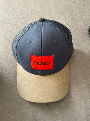 Blå och beige keps från Hugo - Snygg keps från Hugo med mörkblått tyg och mesh på sidorna, beige böjd skärm och röd patch med svart HUGO-logga framtill. Justerbar baktill och ventilerande mesh gör den perfekt för en avslappnad streetwear-look.