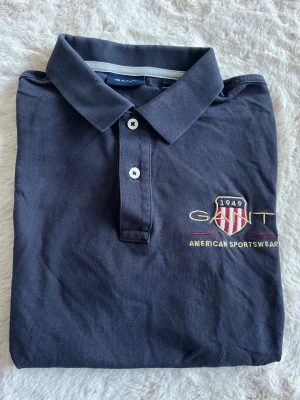 Pikétröja från Gant - Klassisk mörkblå pikétröja från Gant med broderad logga och texten 'American Sportswear' på bröstet. Tröjan har korta ärmar, krage och knappar framtill. 