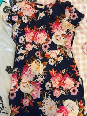 Mörkblå blommig klänning från Lardini - Kortärmad klänning i mörkblått med stora blommiga mönster i rosa, vitt och orange. Klänningen har en markerad midja och rund halsringning. Perfekt för dig som gillar färgstarka och feminina plagg.