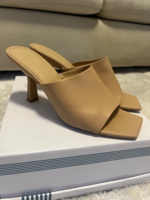 Beige slip-in pumps med fyrkantig tå - Stilrena beige slip-in pumps från H&M med öppen häl och fyrkantig tå. Skorna har en bred rem över foten och en trendig, vinklad klack. Tillverkade i slätt konstläder som ger en modern look.