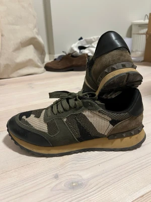Valentino camo sneakers i grönt - Snygga sneakers från Valentino med camomönster i grönt, beige och svart. Skorna har detaljer i mocka och mesh, samt nitar på hälen. Yttersulan är beige och svart, och skorna har gröna skosnören. Perfekta för dig som gillar streetwear och unika detaljer.