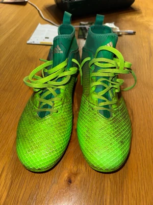 Gröna Adidas fotbollsskor med dobbar - Snygga fotbollsskor från Adidas i en grön färg med skön textur och snörning. Skorna har högre skaft och klassiska dobbar för bra grepp på planen. Tillverkade i syntetmaterial och designade för att sticka ut på fotbollsplanen.