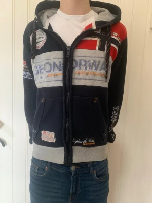 GeoNorway hoodie med tryck och huva - Cool hoodie från GeoNorway med huva och dragkedja. Mörkblå bas med grå och röda detaljer, flera broderade och tryckta loggor samt patchar. Fickor framtill och ribbade muddar. Perfekt för dig som gillar sportig och unik stil.