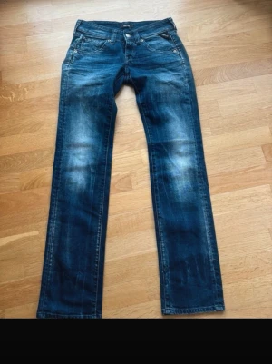 Blå raka jeans från Replay - Säljer ett par klassiska blå jeans med raka ben och snygga slitningar på framsidan. Jeansen har fem fickor, knappgylf och kontrastsömmar. Perfekta för en avslappnad och trendig look. Materialet är denim i bomull. Storlek 25/32 låg midjade