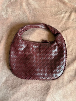 Vinröd flätad handväska från design by sí - Säljer en vinröd handväska med flätad design och rundad form. Väskan har ett kort, vadderat handtag och dragkedja upptill. Tillverkad i skinnimitation som ger en lyxig känsla. Perfekt accessoar för dig som gillar trendiga detaljer. Använd 2 gånger. Har en jätteliten fläck på insidan men inget som påverkar väskan i helheten