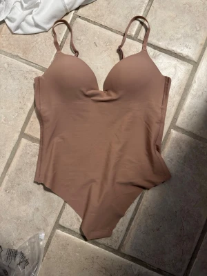 Beige body med vadderade kupor - Snygg beige body med vadderade kupor och justerbara axelband. Tillverkad i ett mjukt, stretchigt material som formar sig efter kroppen. Perfekt som topp eller under tighta plagg för shapewear. Endast testad och var då för stor i bysten på mig som är 70B, syrran på 70C passar den 