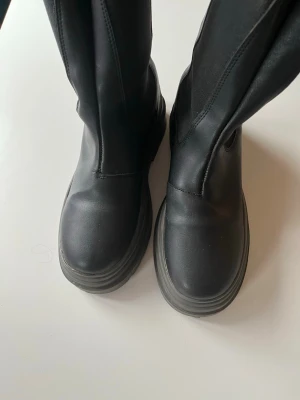 Svarta chunky boots i skinnimitation - Säljer ett par svarta höga boots med chunky sula och dragflik baktill. Stövlarna är i skinnimitation och har en stilren, minimalistisk design utan synliga detaljer. Perfekta för dig som gillar en edgy och modern look.