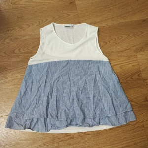 Randig ärmlös linnetopp i blått och vitt - Säljer en luftig och somrig ärmlös linnetopp med vit överdel och blåvit randig nederdel. Toppen har rund halsringning och lätt volang längst ner för en avslappnad look. Perfekt till varma dagar och enkel att matcha.