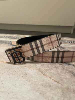  vändbart bälte med TB-spänne - Snyggt vändbart bälte med klassiskt rutigt mönster i beige, svart och rött på ena sidan och diskret svart rutmönster på andra sidan. Stort TB-spänne i metall ger en lyxig touch. Perfekt accessoar för att lyfta din outfit.