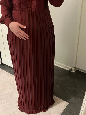 Vinröd långklänning med volangdetaljer - Vinröd långklänning med plisserad kjol, knappar framtill och volangdetaljer på överdelen. Klänningen har långärmade, transparenta ärmar och markerad midja. Elegant och stilren design, perfekt för dig som vill sticka ut.