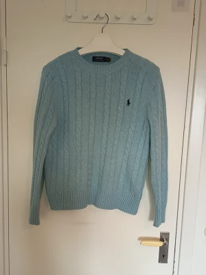 Ljusblå kabelstickad tröja Polo Ralph Lauren - Ljusblå kabelstickad tröja från Polo Ralph Lauren med rund halsringning och klassisk broderad logga på bröstet. Tröjan har ribbade muddar vid ärmslut och nederkant, samt en mjuk och skön känsla. Perfekt för dig som gillar stilrena och tidlösa plagg.