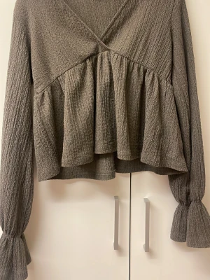 Brun volangblus med lång ärm - Trendig brun  blus med volangdetaljer och struktur i tyget. Blusen har v-ringning och långa ärmar med resår och volang vid ärmslut. Perfekt för dig som gillar en avslappnad men ändå stilsäker look.