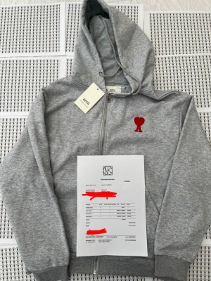Grå zip hoodie från Ami Paris - Säljer en grå hoodie från Ami Paris med dragkedja och huva. På bröstet finns Ami:s ikoniska röda hjärta med bokstaven A broderat. Hoodien har ribbade muddar och är tillverkad i mjuk bomullsblandning. Perfekt för en chill och stilren look.