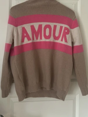 Stickad tröja med AMOUR-tryck - Beige stickad tröja med breda rosa och vita ränder samt texten 'AMOUR' i rosa på ryggen. Tröjan har rund hals och långa ärmar, perfekt för en trendig och avslappnad look.