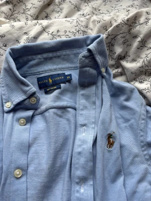 Ljusblå lång kofta från Ralph Lauren - Ljusblå lång kofta från Ralph Lauren i mjuk bomull. Klassisk krage, långa/korta ärmar (går till drygt armbågen) och knappar hela vägen framtill. Man kan använda som en klänning också.  Broderad logga på bröstet för en clean och tidlös stil. Nästan aldrig använt. 