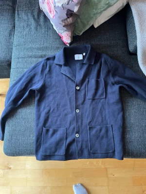 Mörkblå kofta med fickor och krage - Stilren mörkblå kofta från bläck, med tre stora framfickor och klassisk krage. Koftan har knäppning med tre knappar och är tillverkad i ett mjukt, stickat material. Perfekt för lager på lager och enkel att matcha med olika outfits.