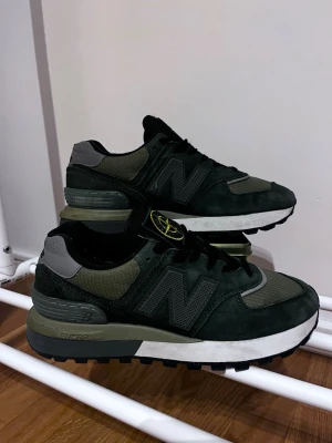 New Balance x Stone Island sneakers - Snygga sneakers från New Balance i samarbete med Stone Island. Skorna har en mörkgrön och olivgrön färgskala med svarta detaljer och en tydlig Stone Island-logga på plösen. Ovandelen är i mocka och mesh, och sulan är vit med svarta och gröna inslag. Perfekta för dig som gillar streetwear.