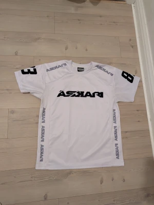 från Askari - Säljer en vit sporttröja från Askari med svart tryck på bröstet och ryggen. Tröjan har siffran 83 på ärmar och rygg samt Askari-logga längs sidorna och axlarna. Tillverkad i ett lätt och ventilerande material, perfekt för träning eller match.