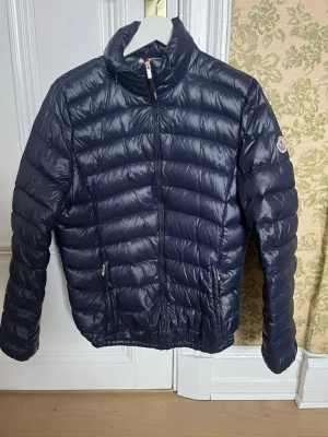 Mörkblå dunjacka från Moncler - Snygg mörkblå dunjacka från Moncler med quiltad design och hög krage. Jackan har dragkedja framtill, två sidofickor med dragkedja och Moncler-logga på ärmen. Tillverkad i glansigt material och fylld med dun för extra värme. Äldre modell. Pris kan diskuteras, och fråga gärna om ni undrar något. 😃