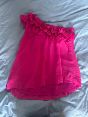 Rosa oneshoulder topp med volang - Säljer en snygg rosa oneshoulder topp med bred volangkant upptill. Toppen har en luftig passform och är tillverkad i ett lätt, syntetiskt material som känns skönt mot huden. Perfekt för dig som vill sticka ut med färg och detaljer.