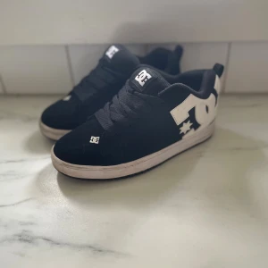 Svarta DC sneakers med vit logga - Säljer ett par klassiska svarta DC sneakers med stor vit DC-logga på sidan och hälen. Skorna har lågt skaft, tjock vit sula och är tillverkade i mockaliknande material med textildetaljer. Perfekta för dig som gillar streetwear och skate-inspirerad stil.