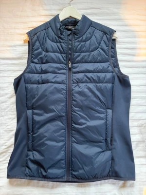 Mörkblå dunväst från Bogeys & Birdies - Snygg mörkblå dunväst från Boomerang med quiltad design och hög krage. Västen har dragkedja framtill och två sidofickor. Tillverkad i ett mjukt, lätt material med dunfyllning för extra värme. Perfekt för lager-på-lager och stilren look.