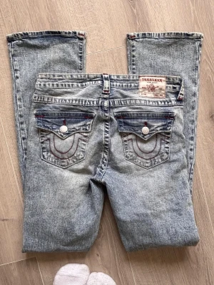True Religion becca jeans - Säljer ett par ljusblå jeans från True Religion med snyggt slitna detaljer och kontrastsömmar. Jeansen har klassiska bakfickor med lock och vita knappar samt röd dekorationssöm. Materialet är denim i bomull och modellen har normal midja.
