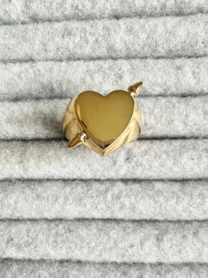 Heart ring - Cool statementring i guldfärgad metall med ett stort hjärta i mitten och två små pilar som sticker ut på sidorna. Ringen har en chunky design och glänsande finish som verkligen drar blickarna till sig. Perfekt för dig som vill ha något unikt och lekfullt i din smyckessamling.