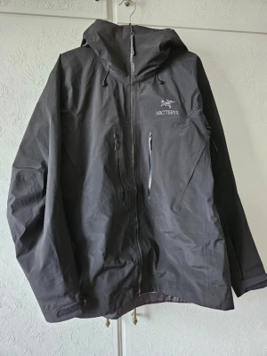 Svart vindjacka från Arc'teryx XXL - Snygg svart vindjacka från Arc'teryx i storlek XXL. Jackan har huva, hel dragkedja framtill och två bröstfickor med dragkedja. Tillverkad i slitstarkt material som står emot vind och lätt regn. Perfekt för utomhusaktiviteter och streetwear.    Jackan är en R3pl1ka
