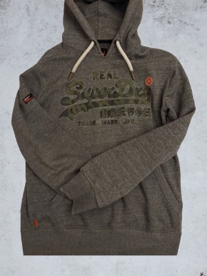 Grå hoodie från Superdry - Säljer en ursnygg grå hoodie från Superdry med snygg text på bröstet! Bara att hojta till för fler bilder! Priset är inte skrivet på sten.