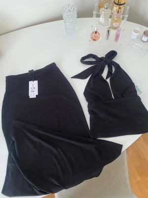 gina tricot set - Snyggt svart set från Gina tricot med mjuk halterneck topp och matchande lång maxikjol. Toppen har knyt i nacken och kjolen är figurnära med stretchigt material. Perfekt för dig som gillar stilrena och bekväma plagg. Materialet är soft touch för extra komfort. Kjolen är i storlek XS och toppen i storlek S