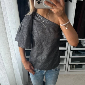Glittrig oneshoulder topp - Snygg glittrig topp i mörkgrått med oneshoulder-design och kort ärm på ena sidan. Perfekt för dig som vill sticka ut med en trendig och unik look. Matcha med jeans eller kjol för en cool outfit. Helt ny med prislapp från Zara strl s