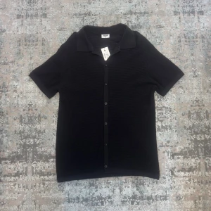 Svart kortärmad blus från Kappahl - Stilren svart blus från Kappahl med korta ärmar och knappar framtill. Blusen har en klassisk krage och är tillverkad i ett mjukt, lätt material med diskret struktur. Perfekt för en clean och enkel look.