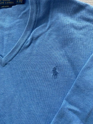 Blå tröja från Polo Ralph Lauren - Snygg blå stickad tröja från Polo Ralph Lauren med V-ringning och diskret broderad logga på bröstet. Tillverkad i mjuk bomull som känns skön mot huden. Mycket bra skick och sparsamt använd. Storlek: L