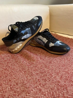 Valentino Rockrunners Blå skor - Snygga sneakers från Valentino med svart, grå och blå camo-mönster. Skorna har svarta skosnören, beige detaljer och coola nitar på hälen. Materialet är en mix av läder och textil, perfekt för dig som gillar att sticka ut med stil.