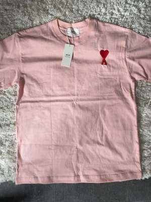 Ami paris t shirt - Säljer en ljusrosa t-shirt från Ami med klassisk rund hals och korta ärmar. På bröstet finns Ami:s ikoniska röda hjärta med bokstaven A broderat. Tillverkad i mjuk bomull för skön känsla och stilren look.